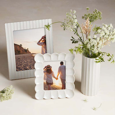 Two white ceramic picture frames and a matching fluted vase with flowers on a neutral surface.